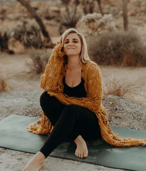 Portrait of Maja Re seated outdoors on a yoga mat, smiling gently, wrapped in a cozy shawl. She is a Breathwork and Yoga Instructor, and Reiki Master.